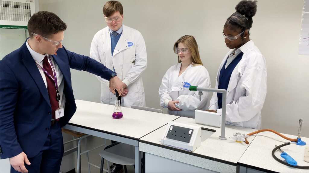 3 students in lab coats with a staff member performing a science experiment